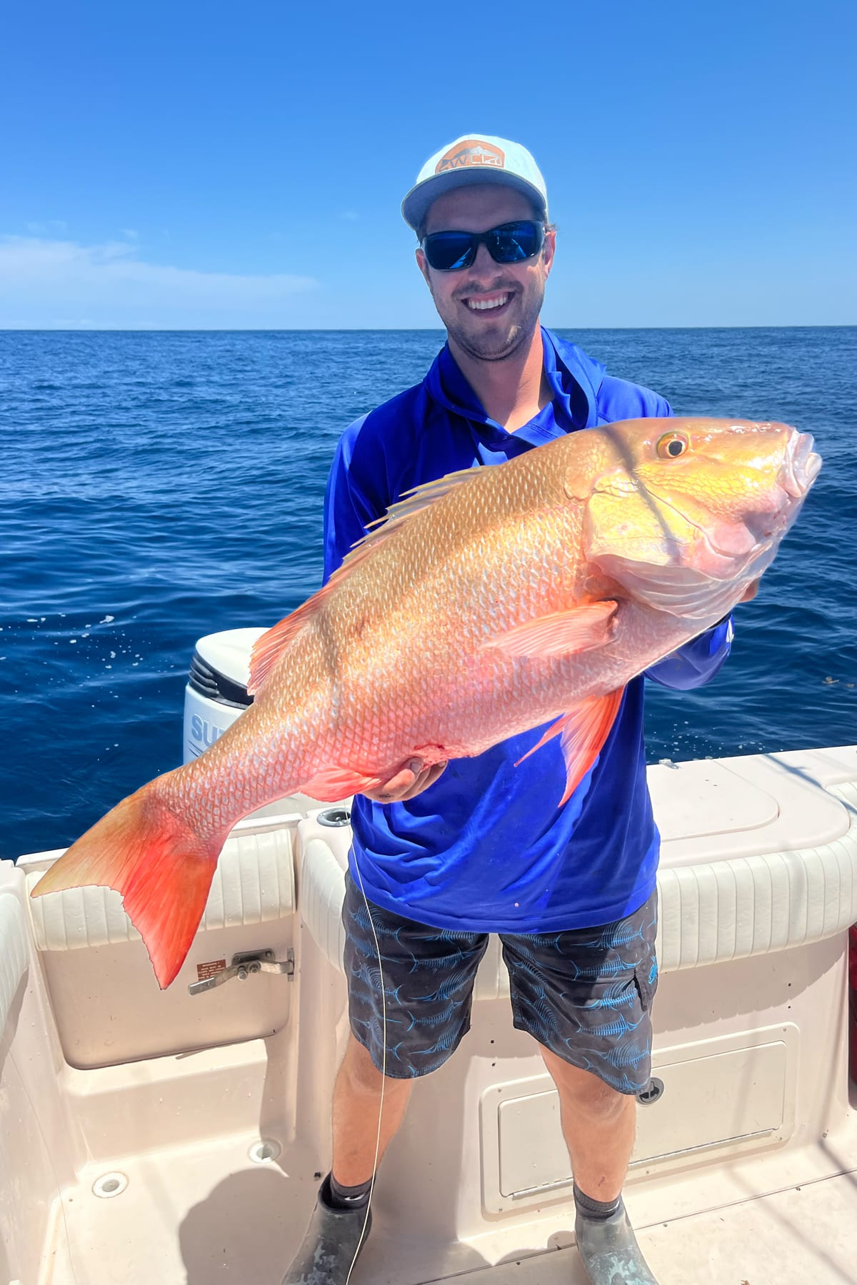 Smiling fisherman on a boat holding a vibrant golden-red mutton snapper against a bright blue ocean backdrop.