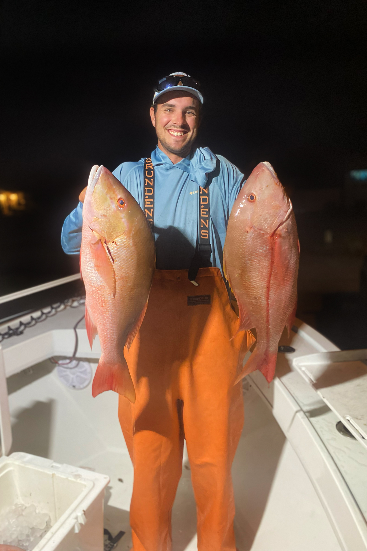  Nighttime catch: fisherman in orange bibs holding two large mutton snapper fish on deck of boat.