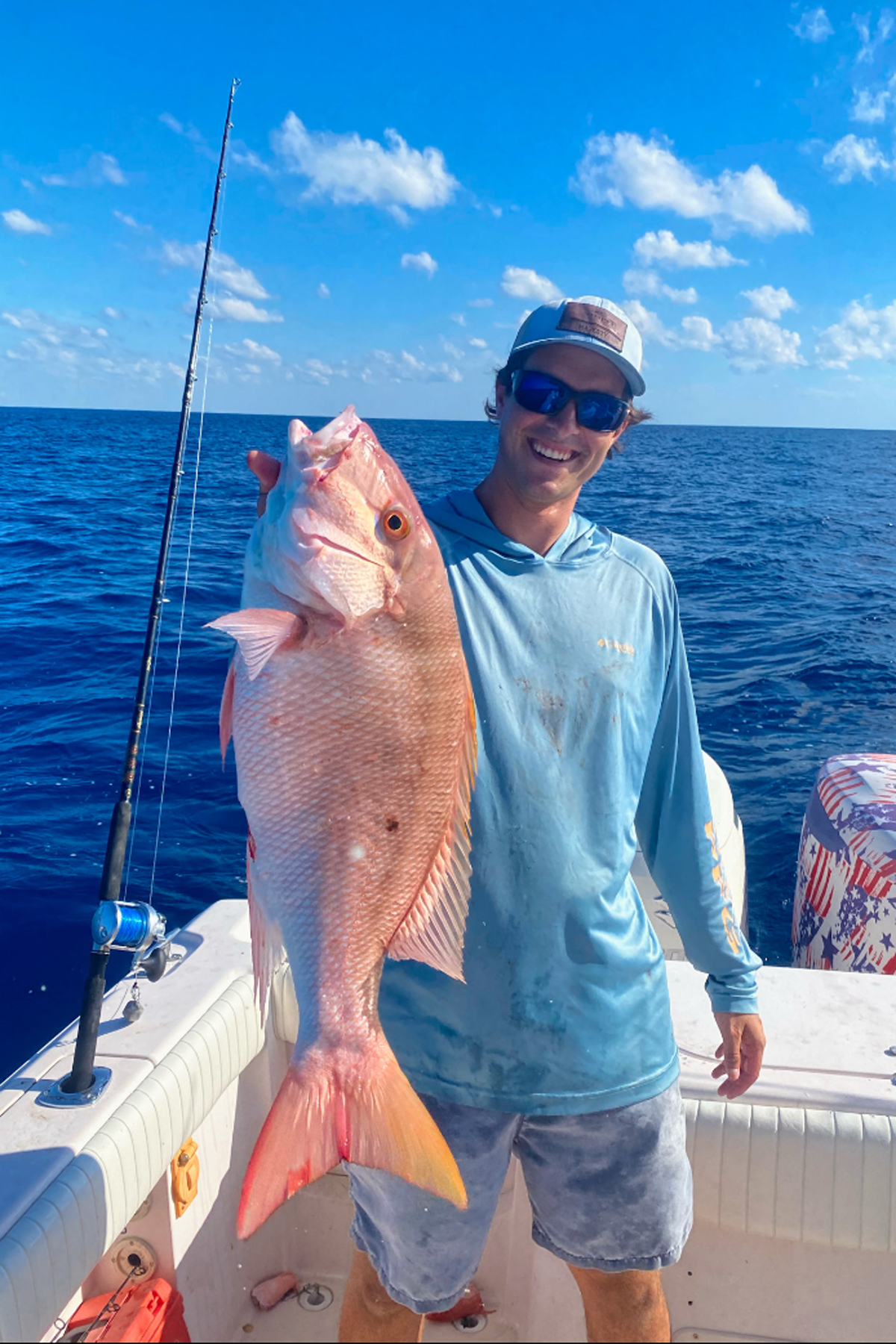  Man wearing sunglasses proudly holding freshly caught mutton snapper on boat, with clear blue sky and deep ocean behind.